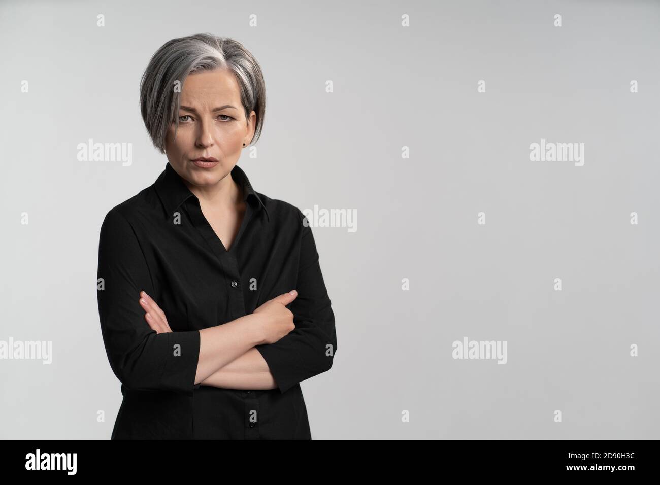 Une femme mûre avec des cheveux gris en chemise noire a plié ses bras sur sa poitrine contre un fond blanc. Espace de copie à droite. Banque D'Images