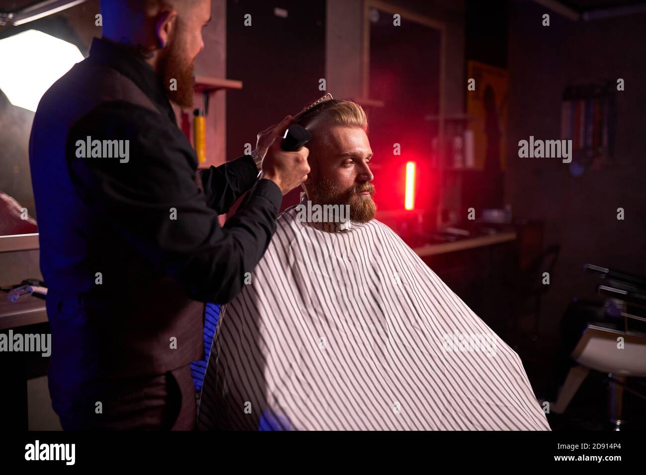 confiant caucasien barber mster coupe les cheveux et la barbe des hommes dans le salon de coiffure, professionnel coiffeur fait la coiffure pour un jeune homme, il travaille avec la tondeuse Banque D'Images