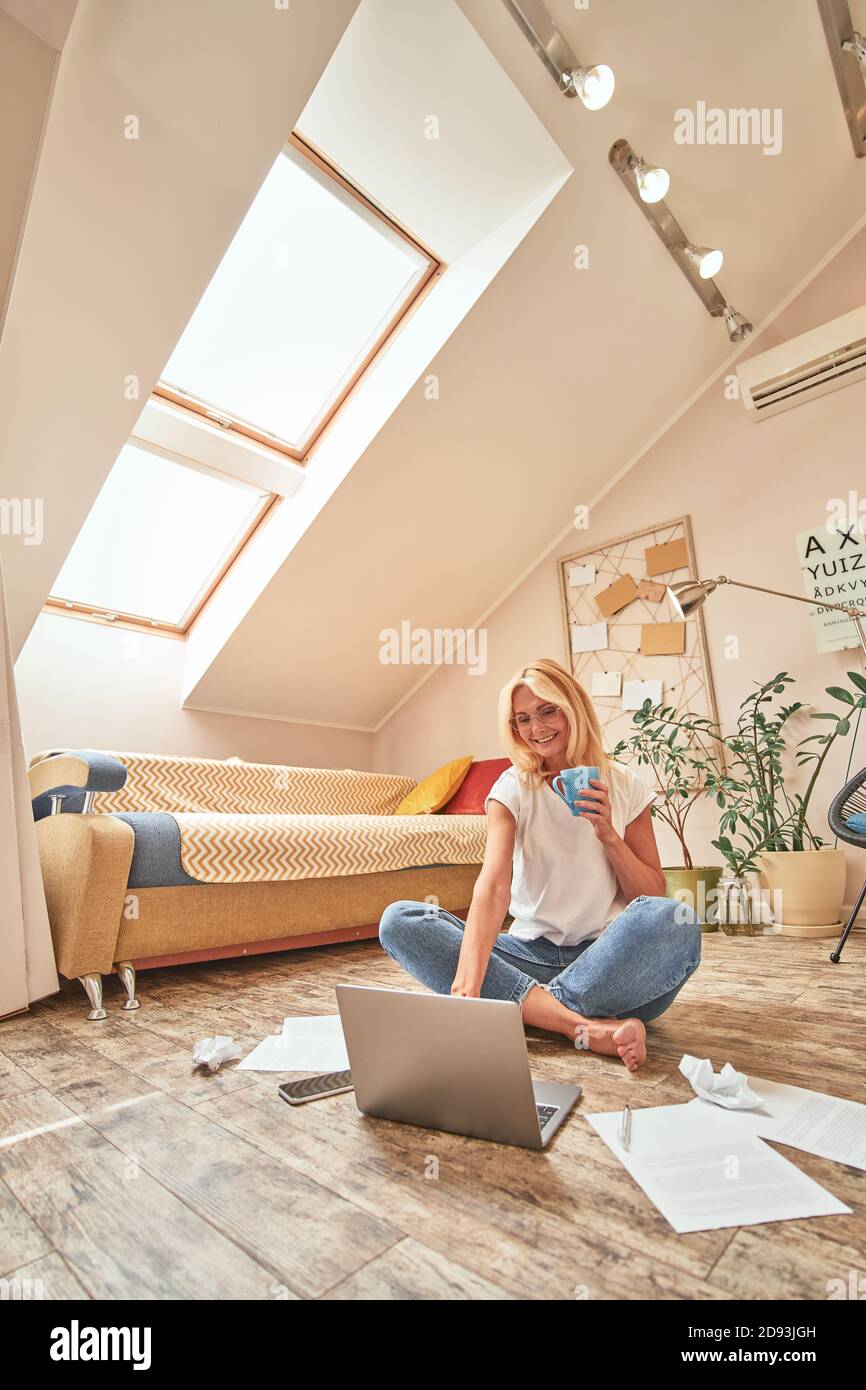 Femme de race blanche d'âge moyen assise sur le sol avec une tasse de café pendant une journée de travail à distance. Travail à domicile et concept indépendant Banque D'Images