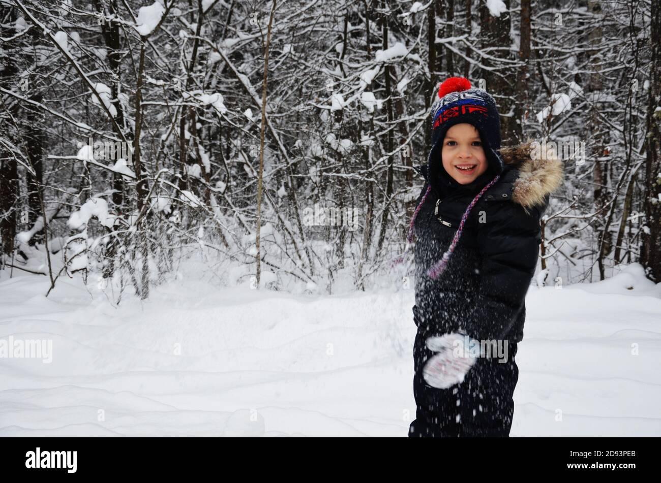 Un petit garçon simpotique jette de la neige dans la forêt enneigée d'hiver, des arbres dans la neige. Aime l'hiver. Joue des boules de neige. Heureux, souriant, riant Banque D'Images