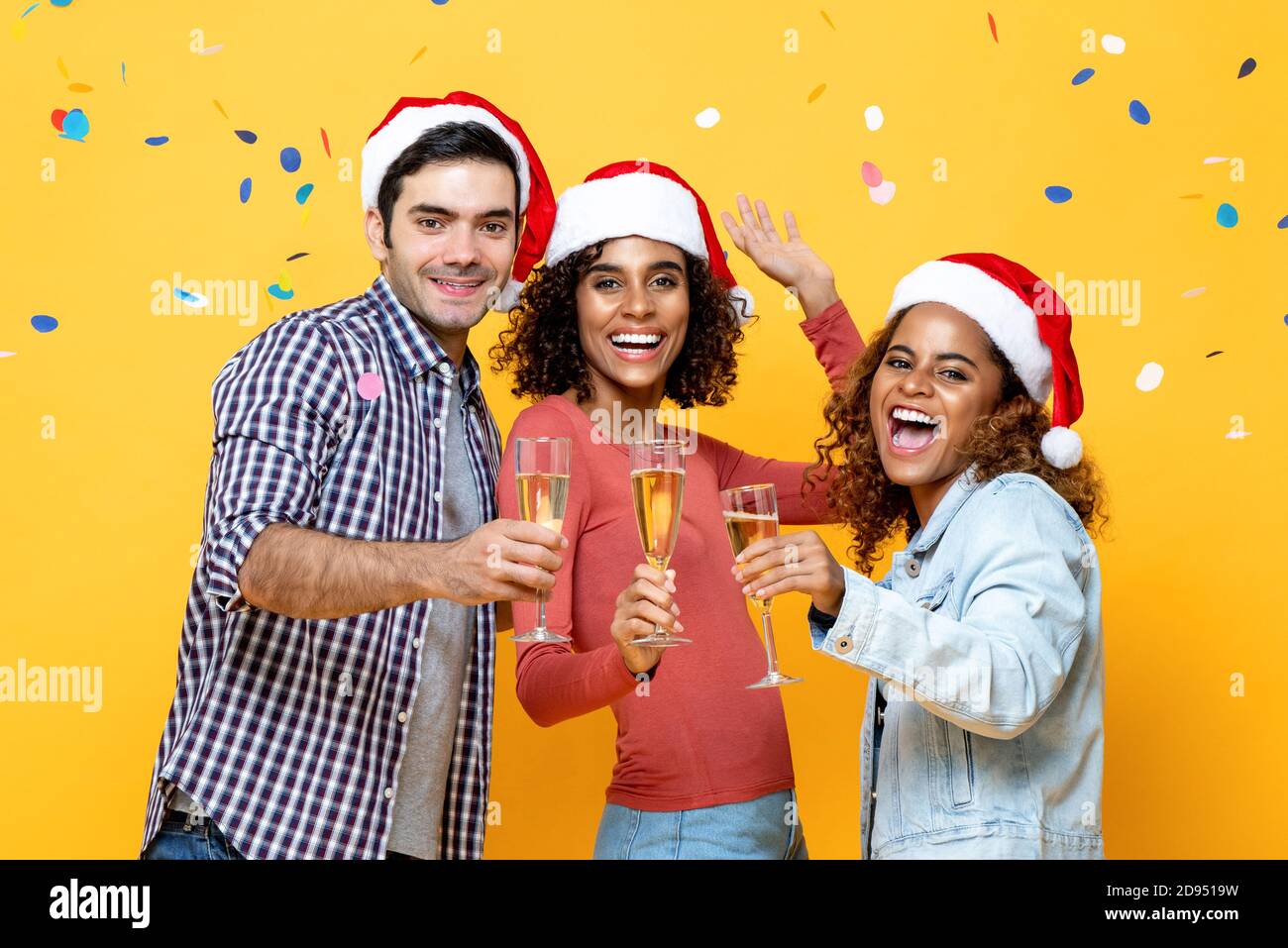 Groupe d'amis multiraciaux heureux célébrant Noël boire du champagne ensemble sur fond jaune studio avec confetti Banque D'Images