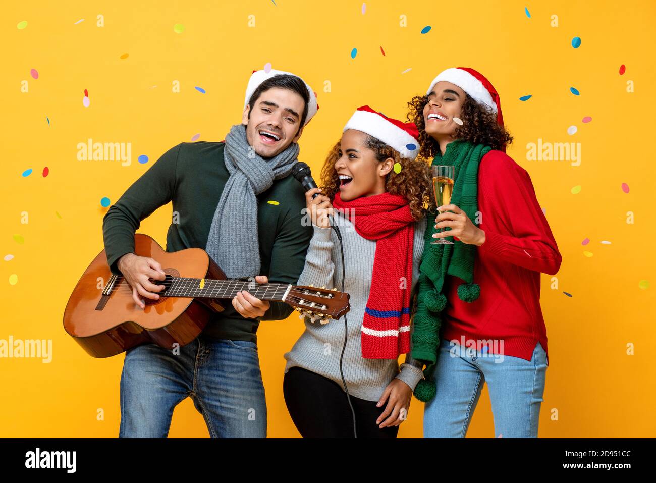 Trois heureux amis multiraciaux chantant ensemble dans la fête de Noël sur fond jaune studio avec confetti Banque D'Images