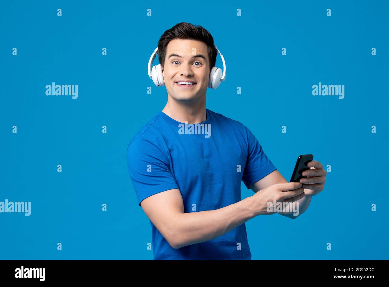 Portrait d'un homme caucasien surpris portant un casque à l'écoute en ligne diffusion de musique sur un téléphone portable isolé sur fond de studio bleu Banque D'Images
