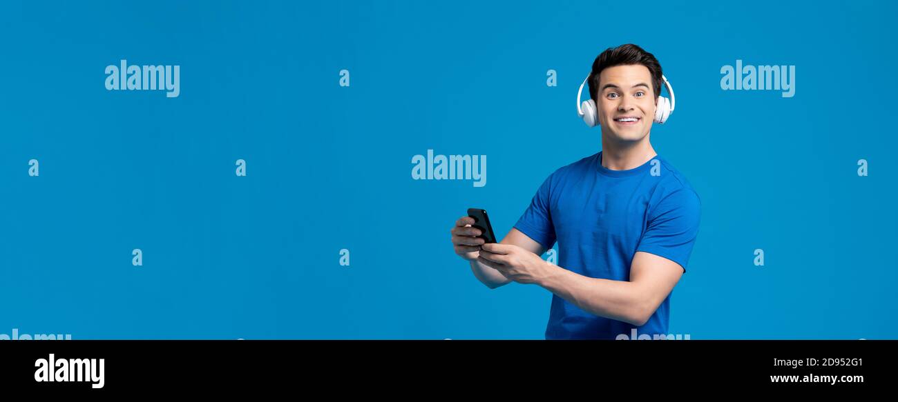 Portrait panoramique d'un homme caucasien surpris portant un casque à l'écoute diffusion de musique en ligne sur un téléphone portable, isolée sur un écran bleu bannière studio Banque D'Images