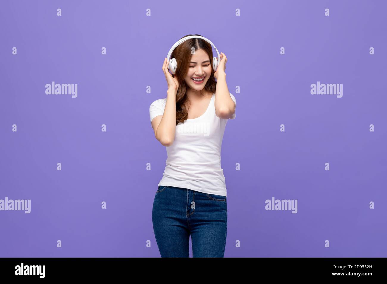Bonne femme asiatique souriante portant un casque pour écouter de la musique avec yeux fermés sur le fond violet isolé Banque D'Images