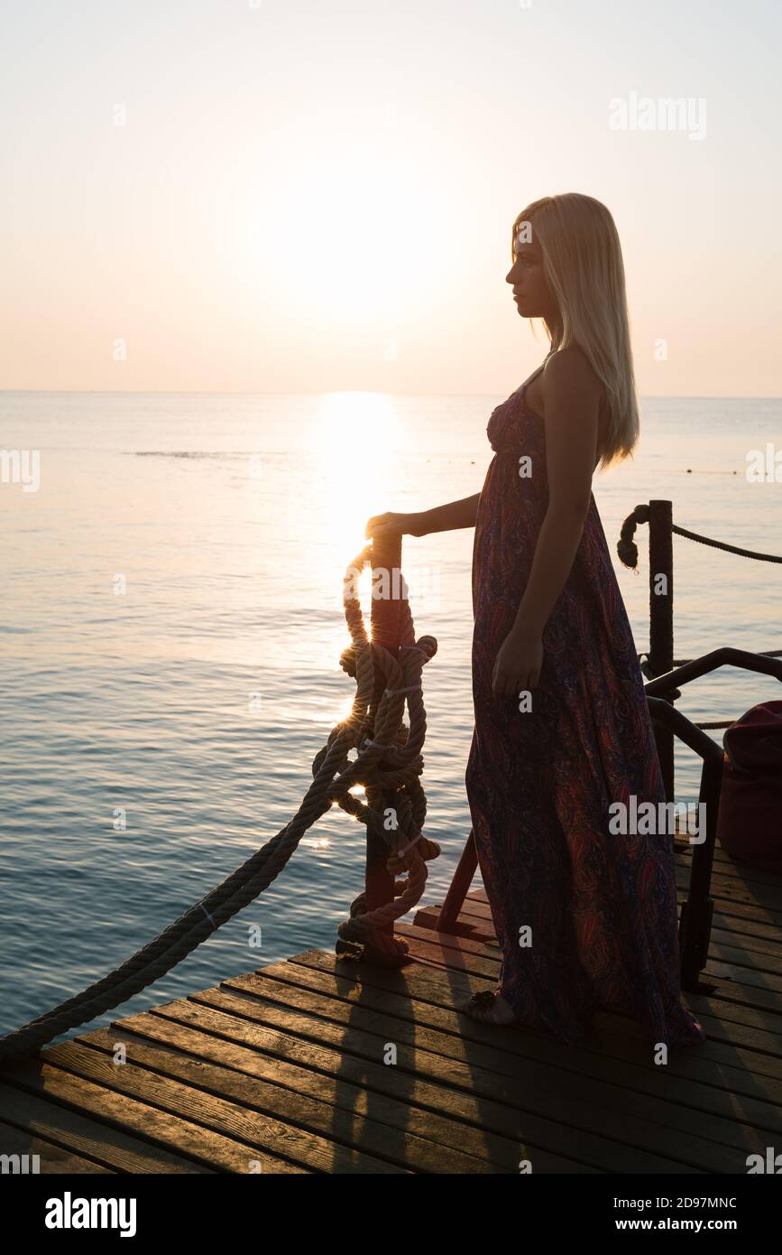 Femme blonde dans une longue sundress marche au bord de la mer à l'aube Banque D'Images