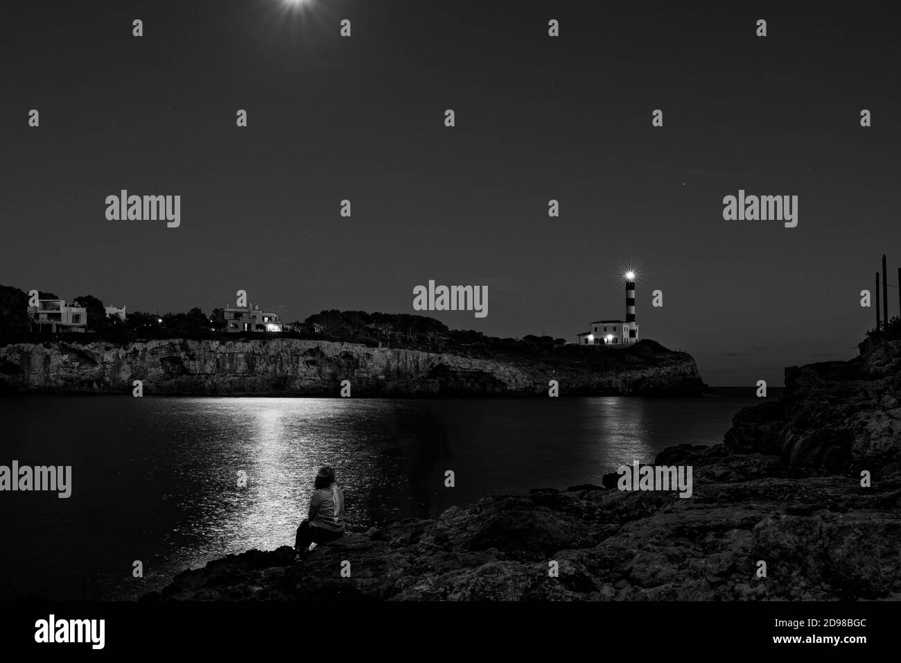 jeune femme assise en face de l'océan avec un reflet au clair de lune et un phare la nuit. image poétique et relaxante, pleine d'esprit, à porto colom Banque D'Images