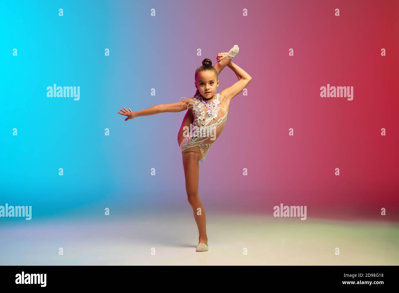 Énergie. Petite fille caucasienne, formation de gymnaste rhytmique, exécutant isolé sur fond dégradé bleu-rouge studio au néon. Enfant gracieux et souple, fort. Concept de sport, de mouvement, d'action. Banque D'Images