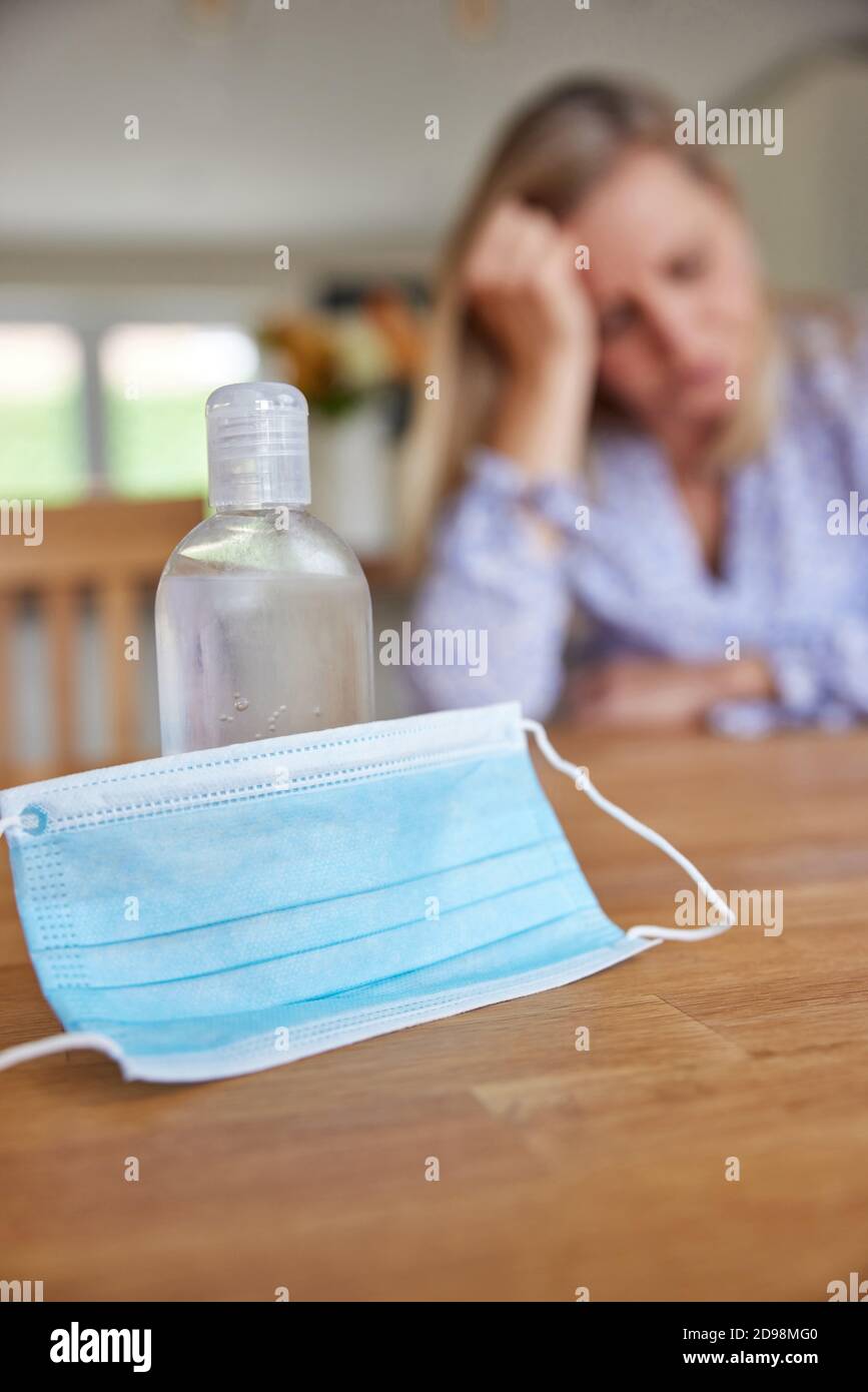 Femme dépressive assise à la maison pendant une pandémie avec masque facial Et bouteille d'assainisseur dans le premier plan Banque D'Images