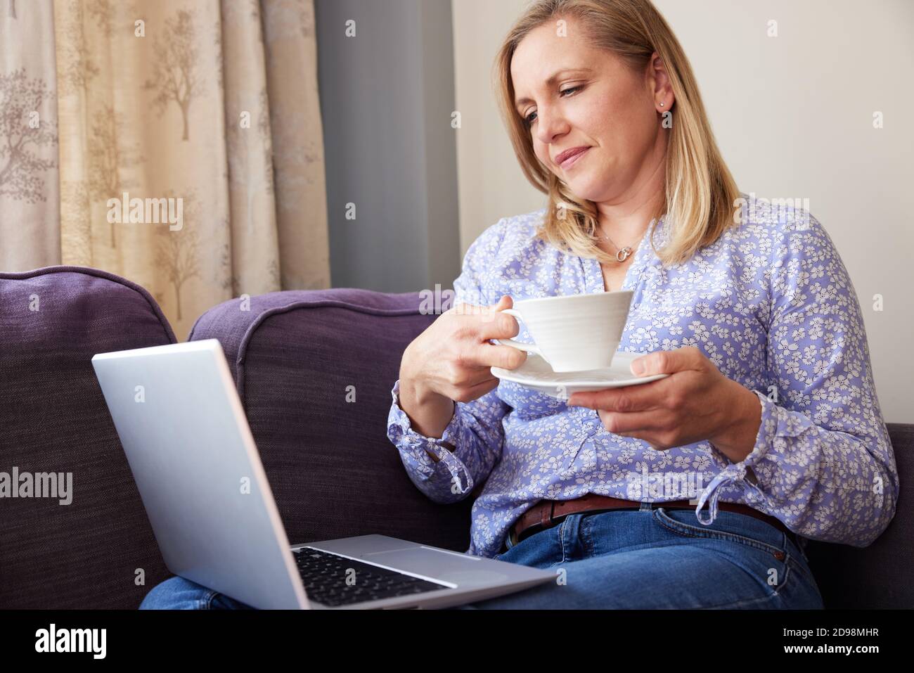 Femme mûre assise sur un canapé regarder l'ordinateur portable et boire Boisson chaude Banque D'Images