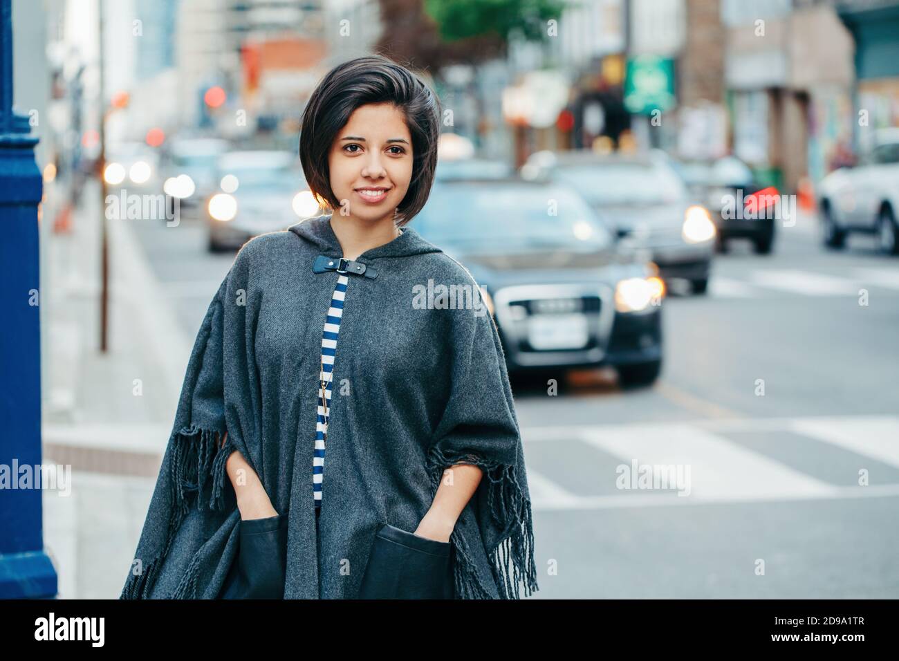 Belle latine jeune femme en gris élégant poncho avec frange dans la rue urbaine. Mode et style urbain. Jolie femme hispanique brune Banque D'Images