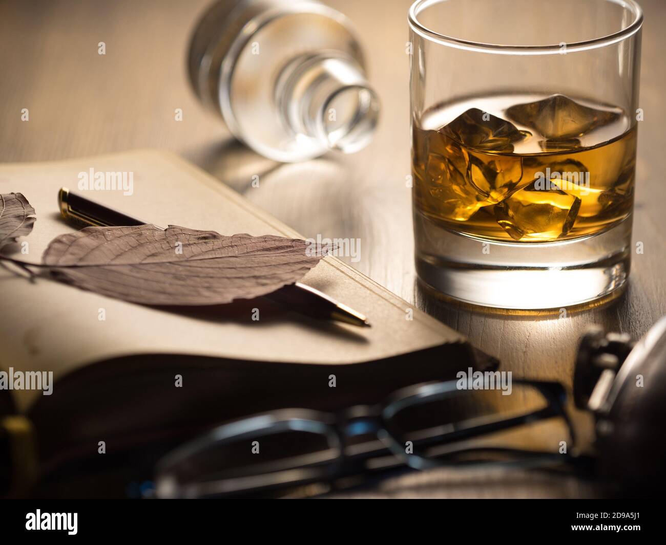 Verre de whisky rétroéclairé sur les rochers, sur une table en bois, avec carnet, stylo et lunettes de lecture Banque D'Images