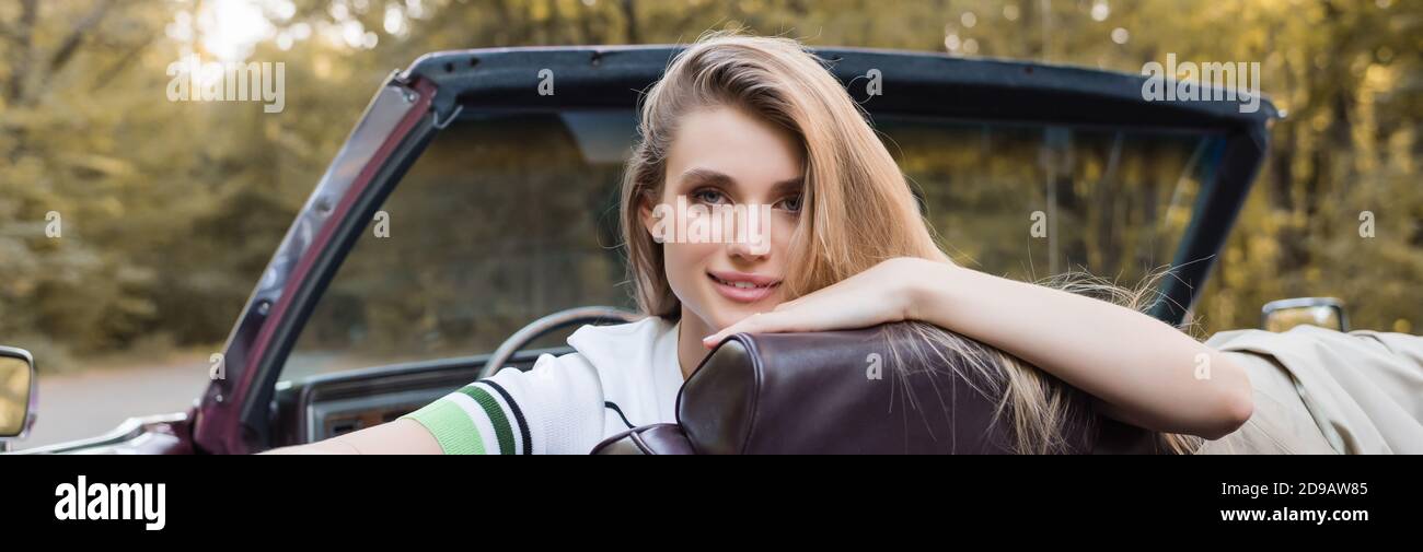 jeune femme souriante regardant l'appareil photo tout en étant assise dans une voiture convertible, bannière Banque D'Images