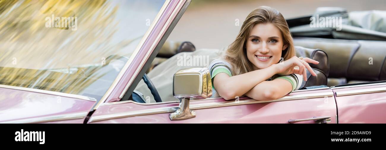jeune femme gaie souriant à la caméra tout en étant assise dans une voiture convertible, bannière Banque D'Images