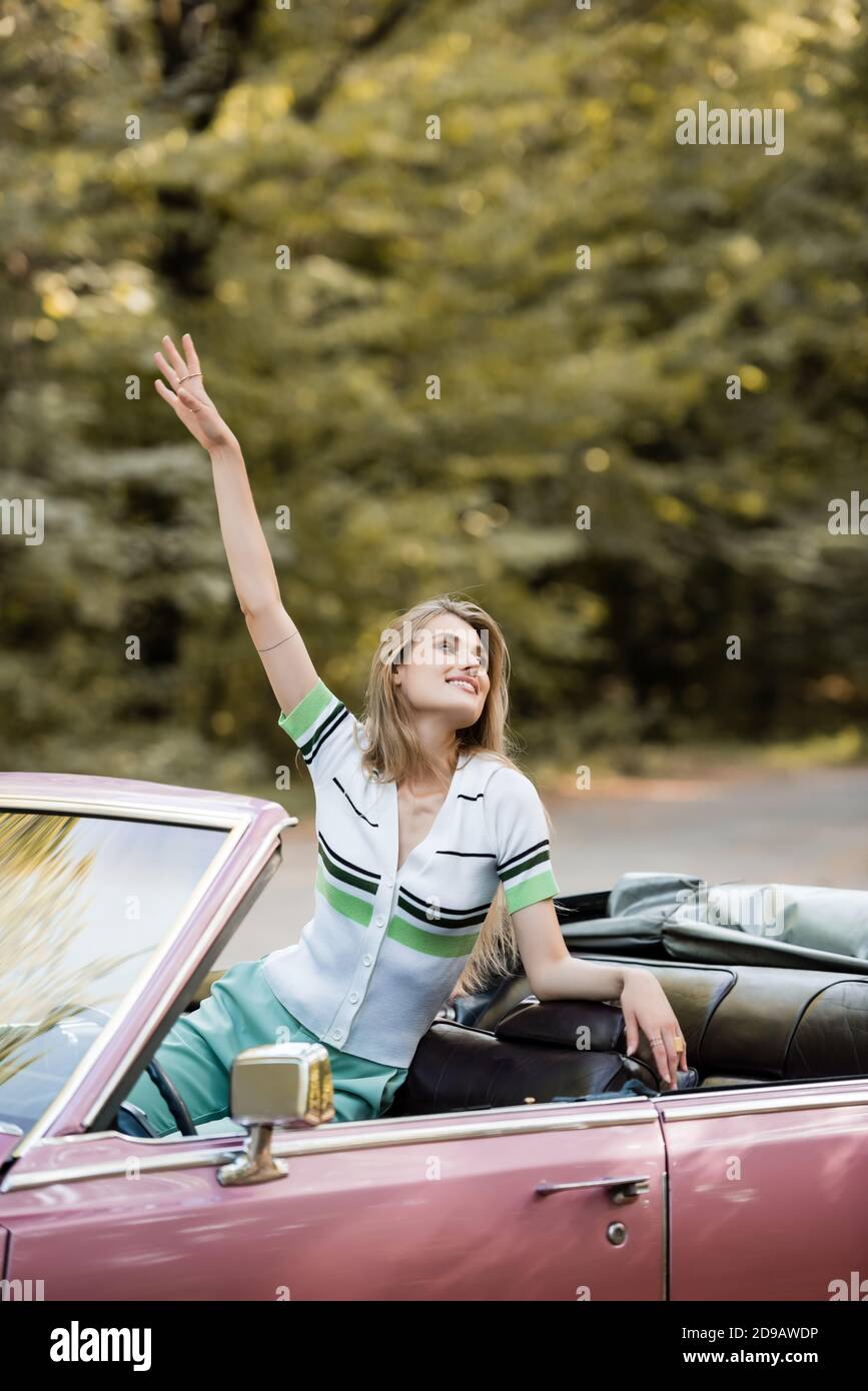 femme souriante regardant loin en étant assise dans une voiture convertible avec main dans l'air Banque D'Images