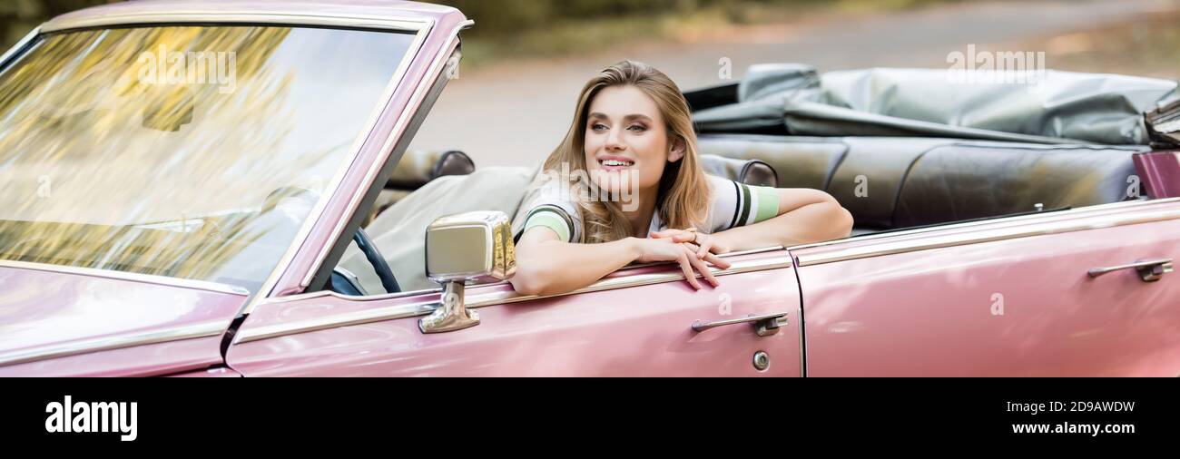 jeune femme joyeuse regardant loin en étant assise dans une voiture convertible, bannière Banque D'Images