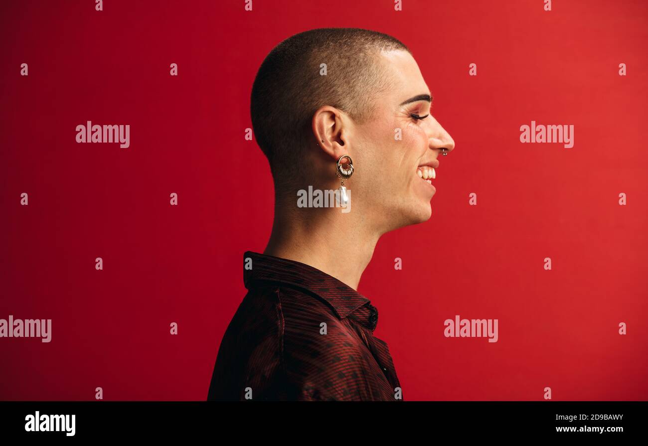 Vue du profil de l'homme androgyne portant un contour d'oreille et un maquillage souriant. Homme gay souriant sur fond rouge. Banque D'Images