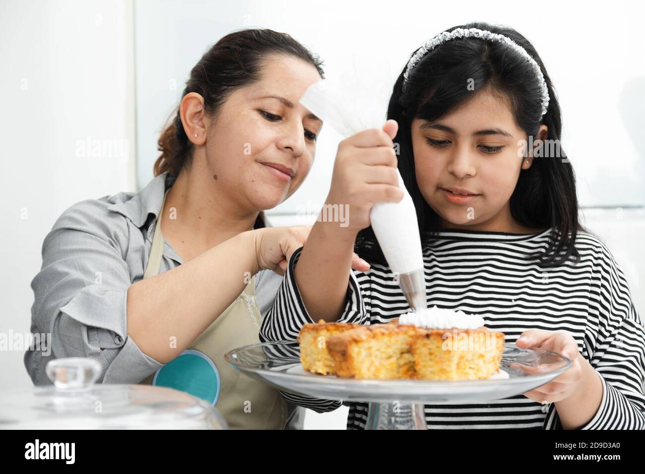 Maman et fille hispanique décorant un gâteau avec de la crème fouettée - maman latine enseignant à sa fille de décorer un gâteau Banque D'Images