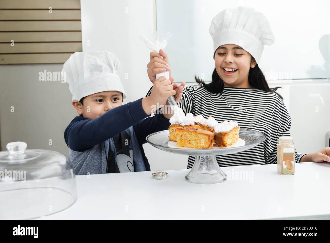 Enfants hispaniques décorant un gâteau avec de la crème fouettée - Latino frères et sœurs s'amuser tout en mettant de la crème fouettée sur le gâteau - les enfants cuisent Banque D'Images