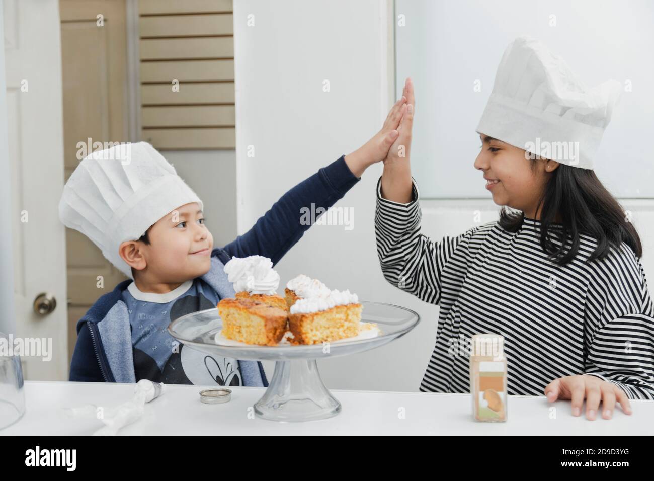 Frères et sœurs latins en chapeaux de chef donnant cinq - enfants hispaniques s'amuser tout en mettant de la crème fouettée sur le gâteau - enfants cuisson Banque D'Images