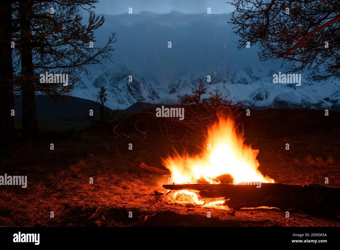 Vue de nuit avec une belle flamme de feu de camp orange et des étincelles contre le fond de montagnes enneigées, de fermes et de ciel nuageux, tourné à longue exposition Banque D'Images