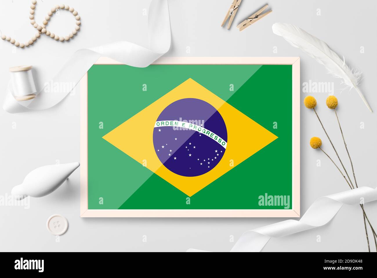 Drapeau brésilien dans un cadre en bois sur fond blanc créatif. Thème blanc, plumes, Marguerite, bouton, objets ruban. Banque D'Images