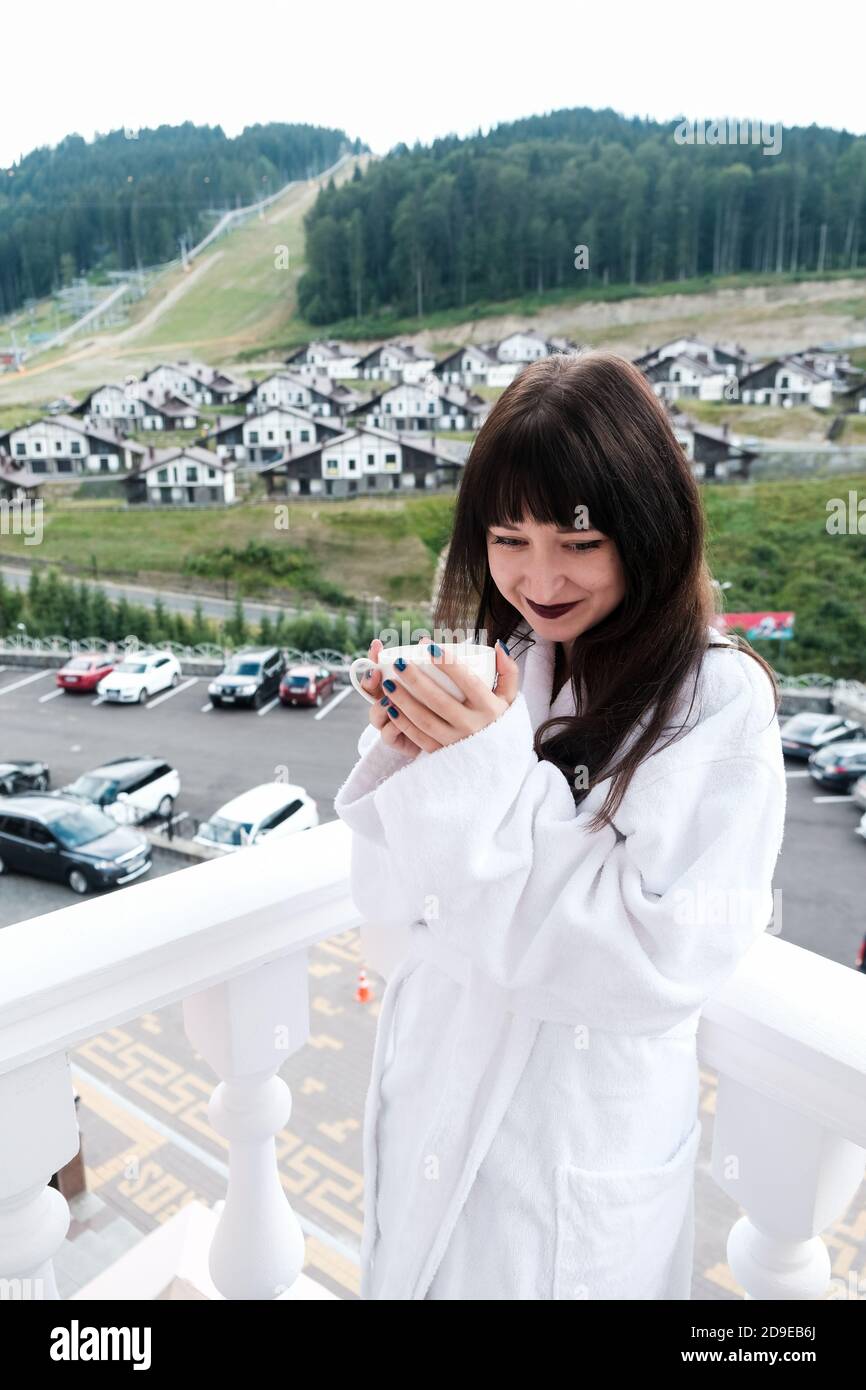 Belle fille timide dans un hôtel blanc peignoir de boire du café chaud sur un balcon. Nature et belle vue à l'extérieur. Escapade tranquille en montagne. Hol Banque D'Images