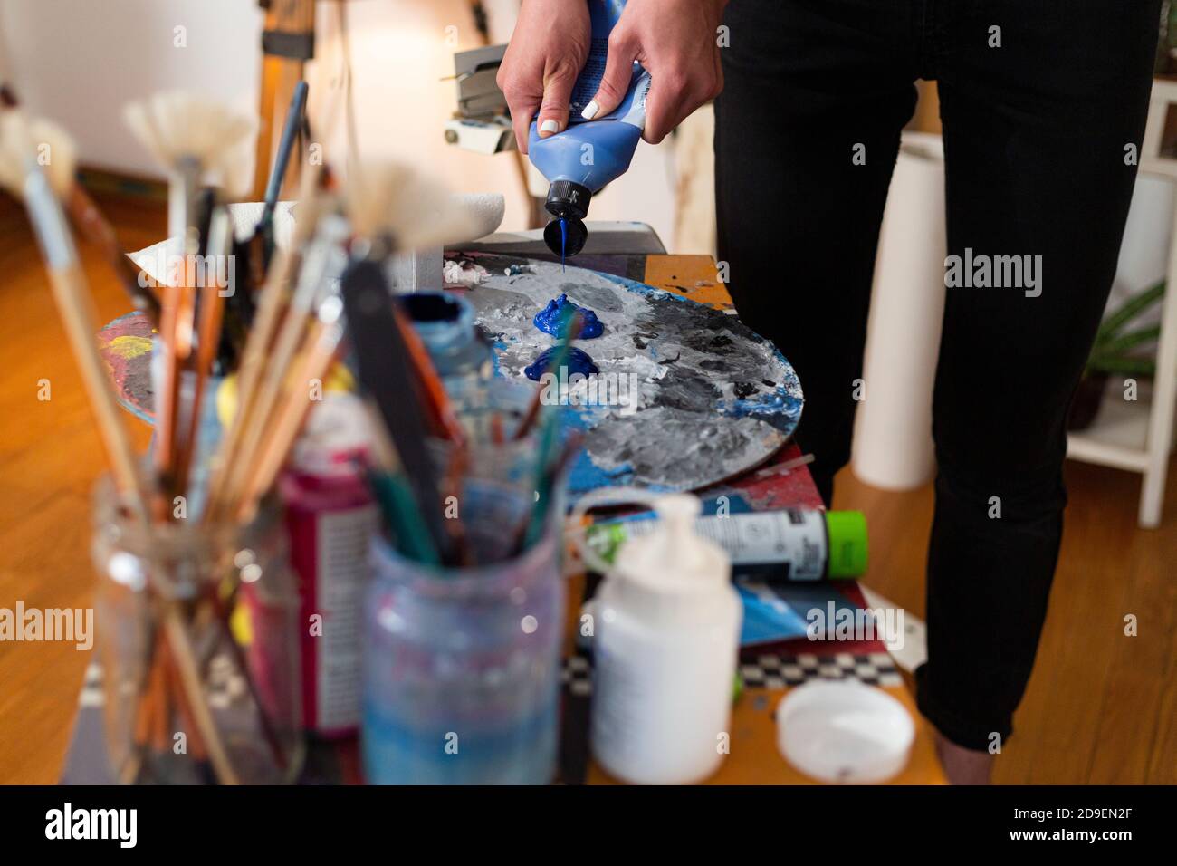 Gros plan de la main du peintre extrudant de la couleur sur une palette dans son studio. Autour de la palette se trouvent une multitude de pinceaux pour peindre en pots. Banque D'Images