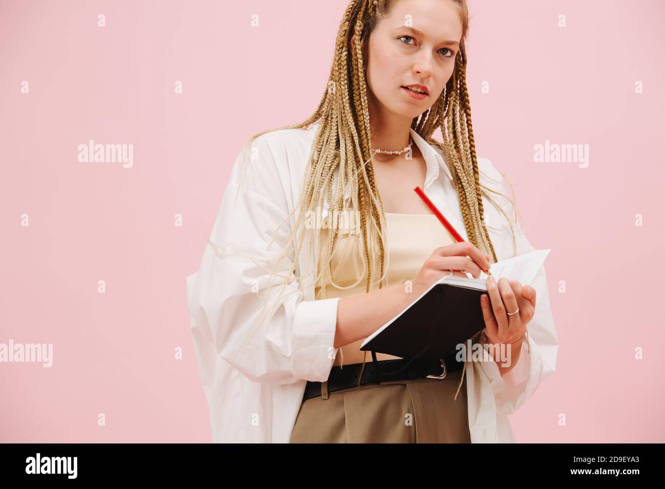 Jeune femme enthousiaste avec des tresses afro blondes élégantes un carnet sur rose Banque D'Images