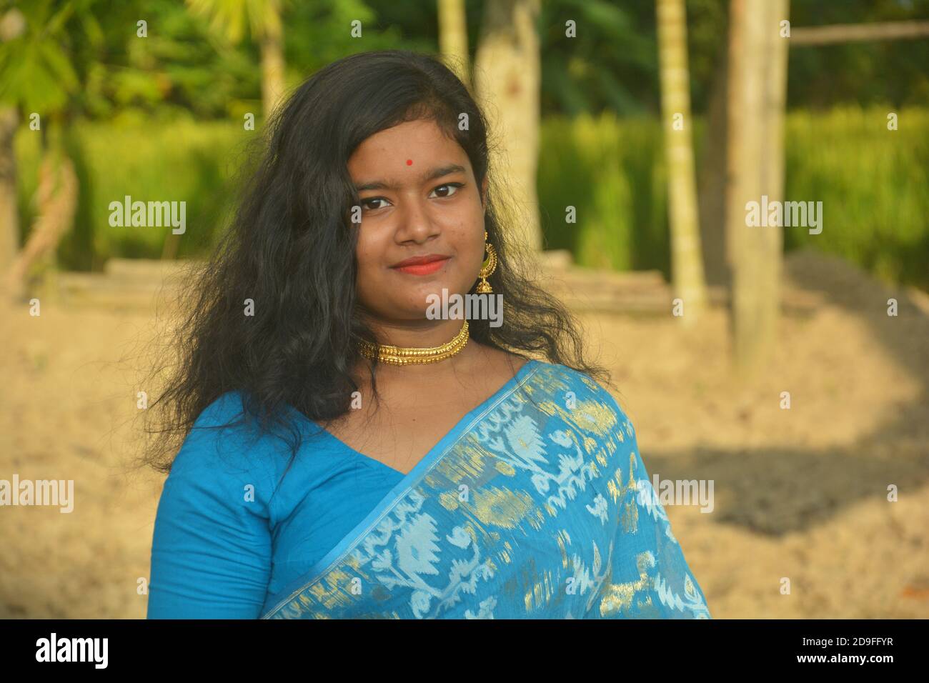 Gros plan d'une jeune fille bengali indienne portant un sari bleu avec des cheveux longs et une pointe de nez dorée, boucles d'oreilles, collier dans un champ labouré, foyer sélectif Banque D'Images