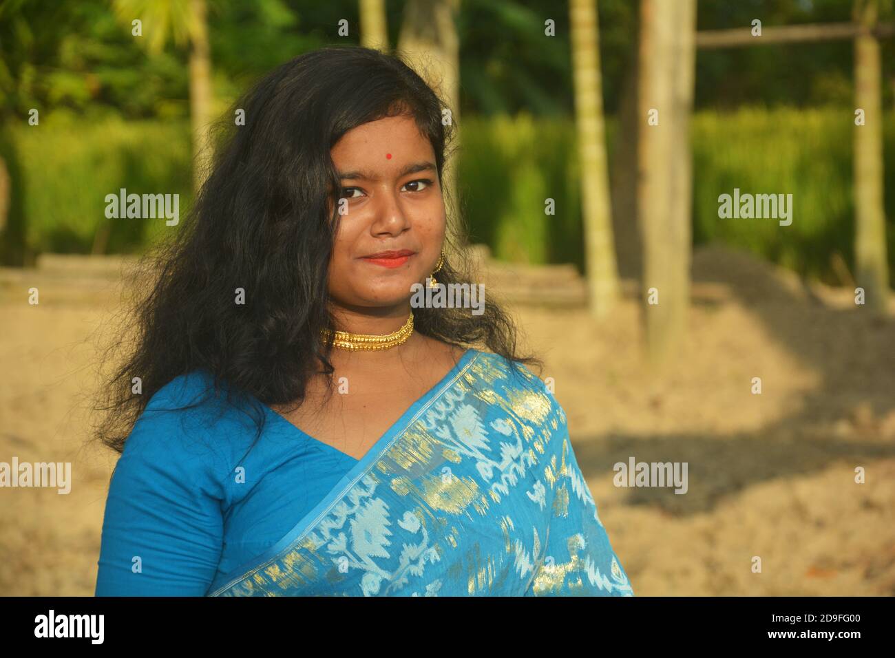Gros plan d'une jeune fille bengali indienne portant un sari bleu avec des cheveux longs et une pointe de nez dorée, boucles d'oreilles, collier dans un champ labouré, foyer sélectif Banque D'Images