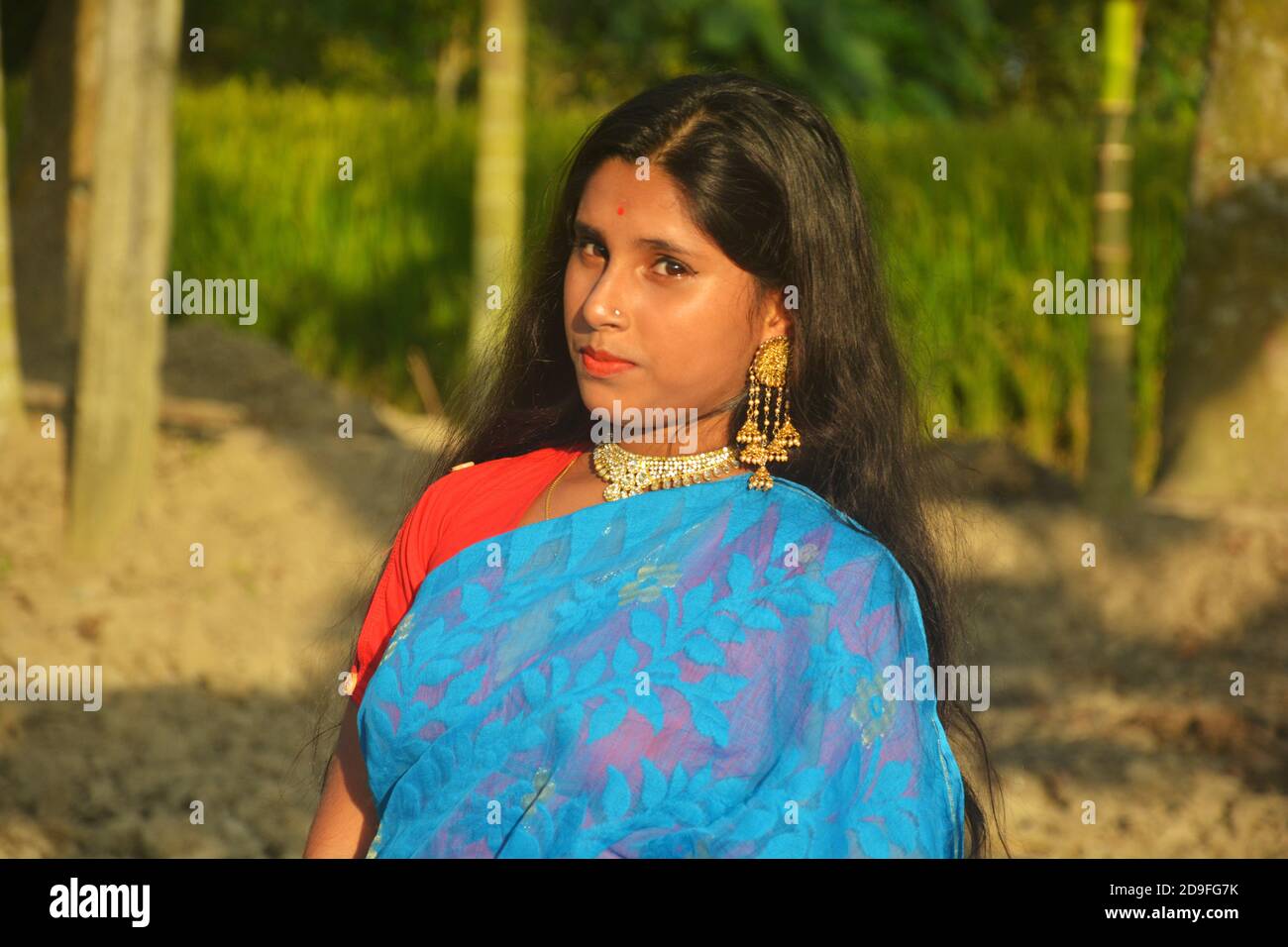 Gros plan d'une jeune fille bengali indienne, portant un sari bleu avec des cheveux longs et une pointe de nez dorée, des boucles d'oreilles, un collier dans un champ labouré, un foyer sélectif Banque D'Images