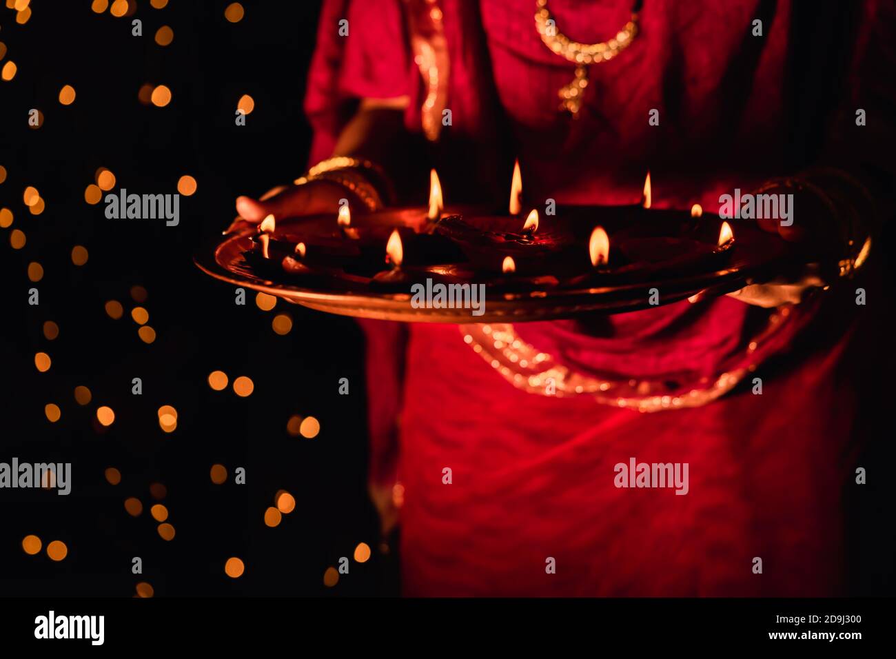 Femme indienne ou mariée portant une robe rouge traditionnelle et des bijoux, tenant puja thaali plein de lampes diya ou argile illuminées, célébrant Diwali la nuit. Banque D'Images