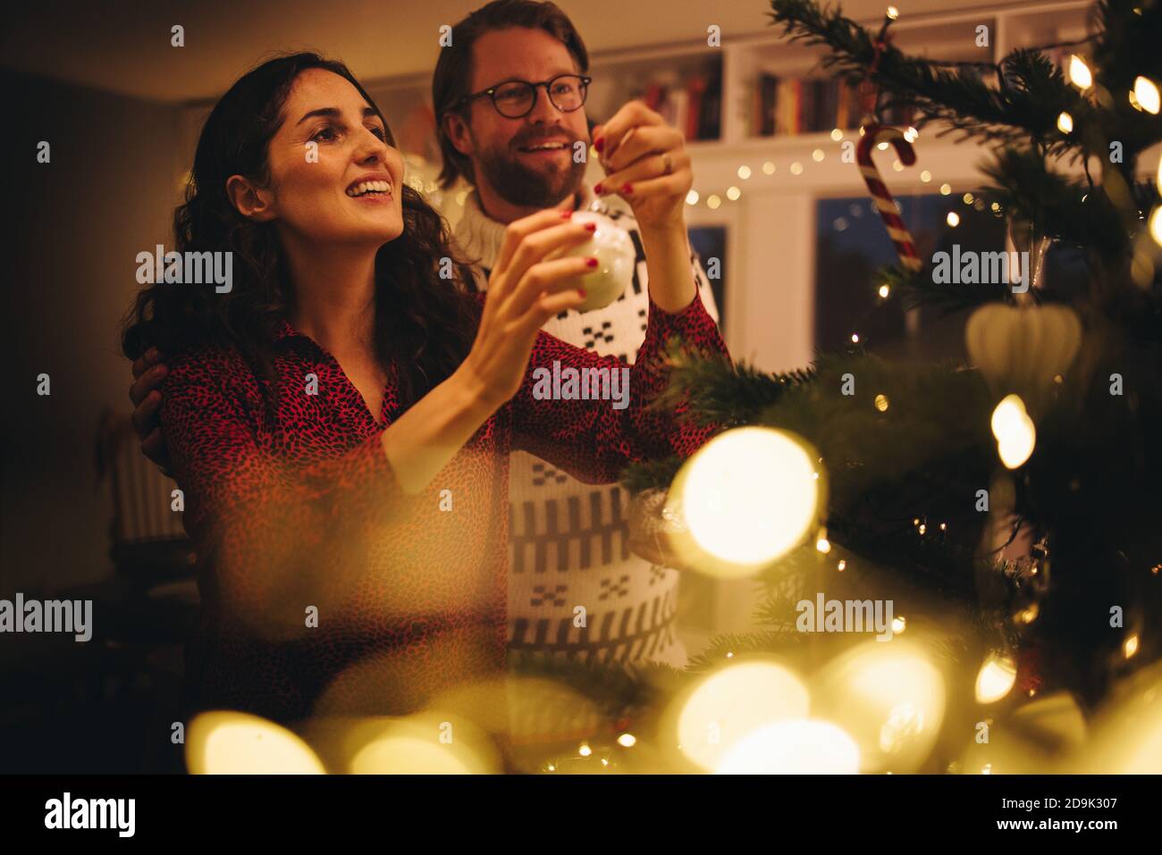 Couple décorant arbre de Noël à la maison de famille. Femme nouant une boule décorative brillante sur l'arbre de Noël avec un homme debout près. Banque D'Images