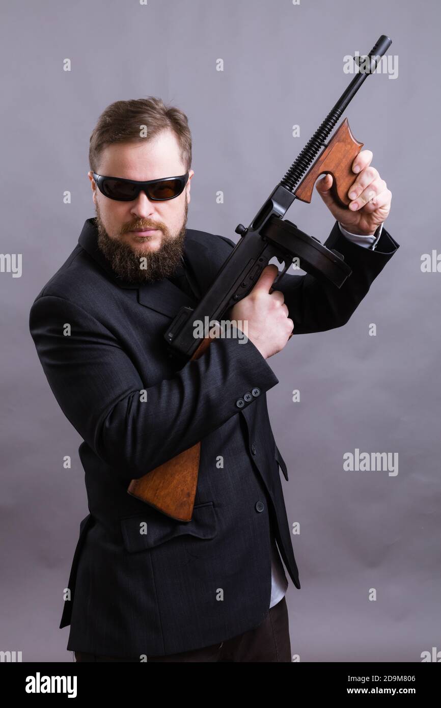 Homme à barbe mature en lunettes de soleil vêtu en costume avec tommy canon sur fond gris Banque D'Images