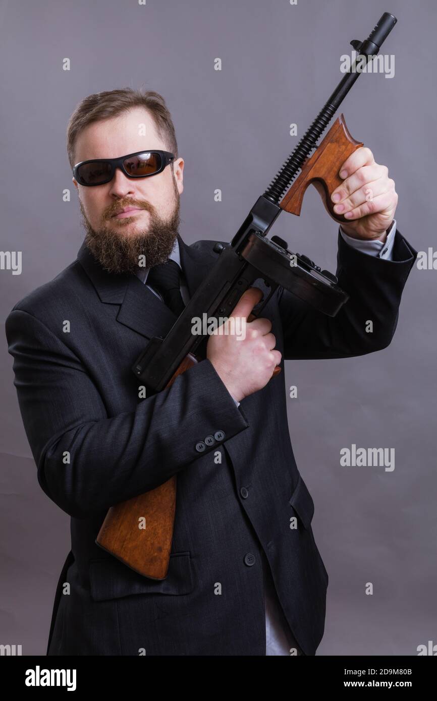 Homme à barbe mature en lunettes de soleil vêtu en costume avec tommy canon sur fond gris Banque D'Images