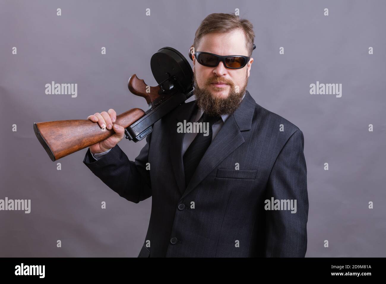 Homme à barbe mature en lunettes de soleil vêtu en costume avec tommy canon sur fond gris Banque D'Images