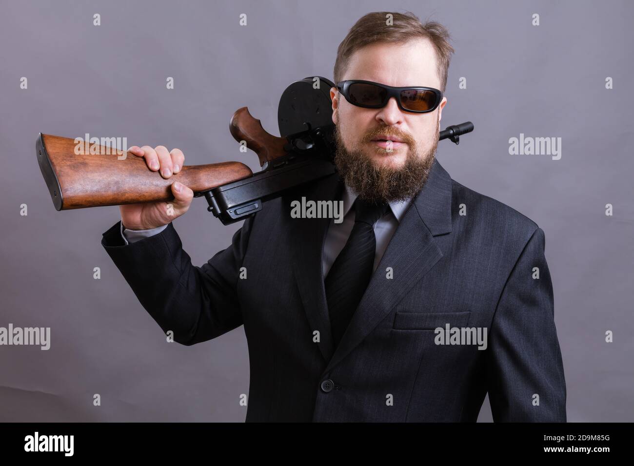 Homme à barbe mature en lunettes de soleil vêtu en costume avec tommy canon sur fond gris Banque D'Images