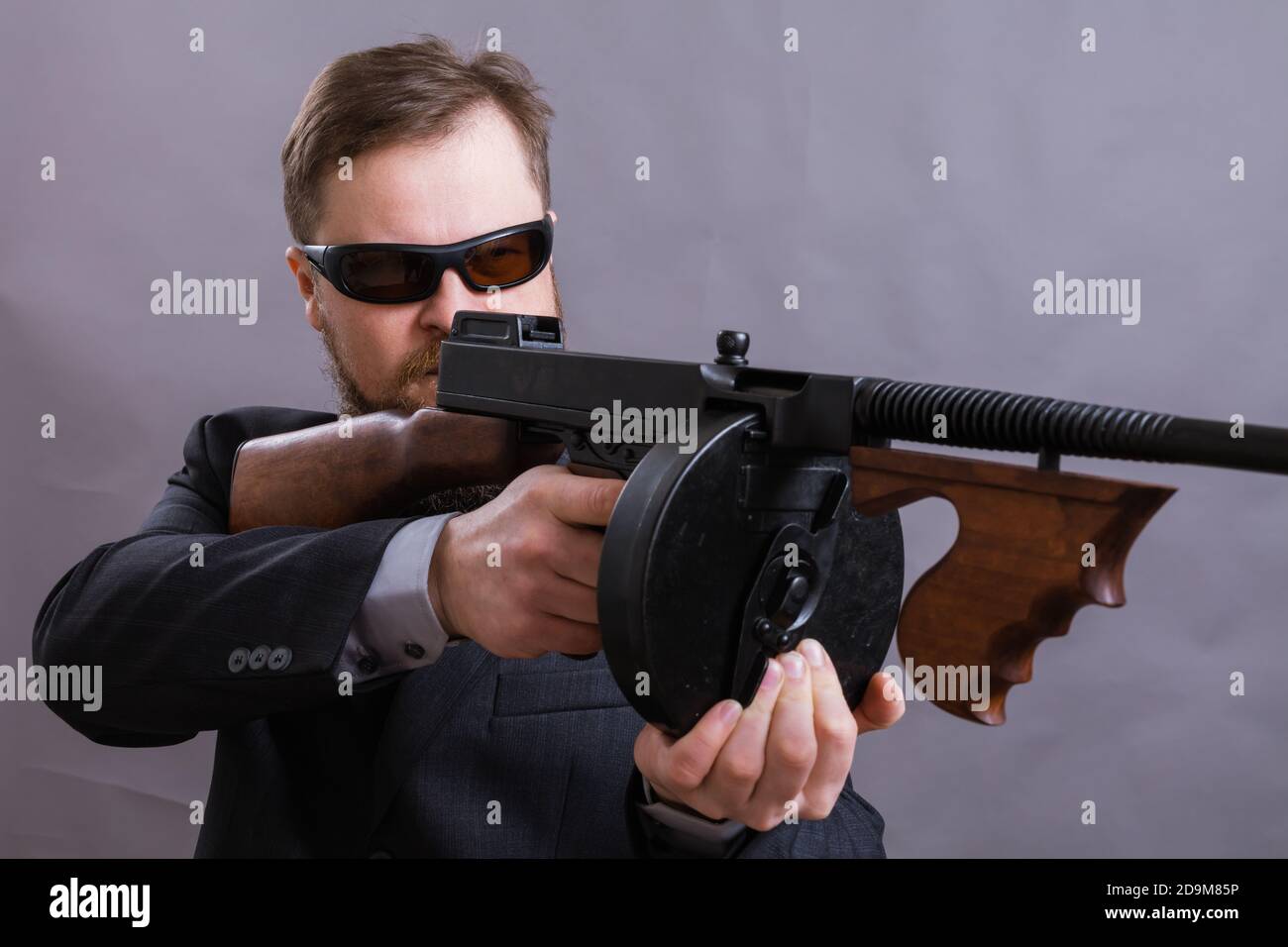 Homme à barbe mature en lunettes de soleil vêtu en costume avec tommy canon sur fond gris Banque D'Images