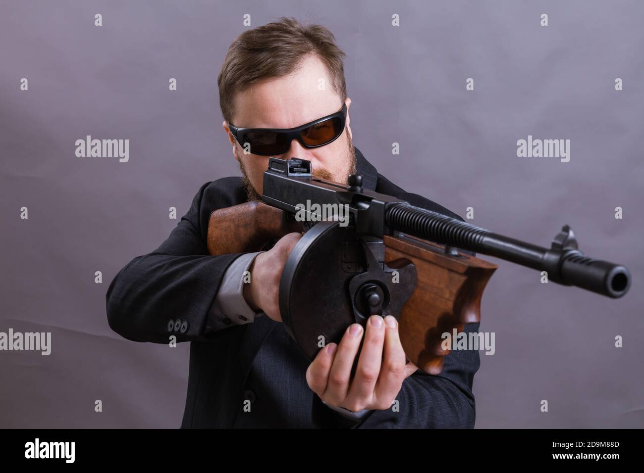 Homme à barbe mature en lunettes de soleil vêtu en costume avec tommy canon sur fond gris Banque D'Images