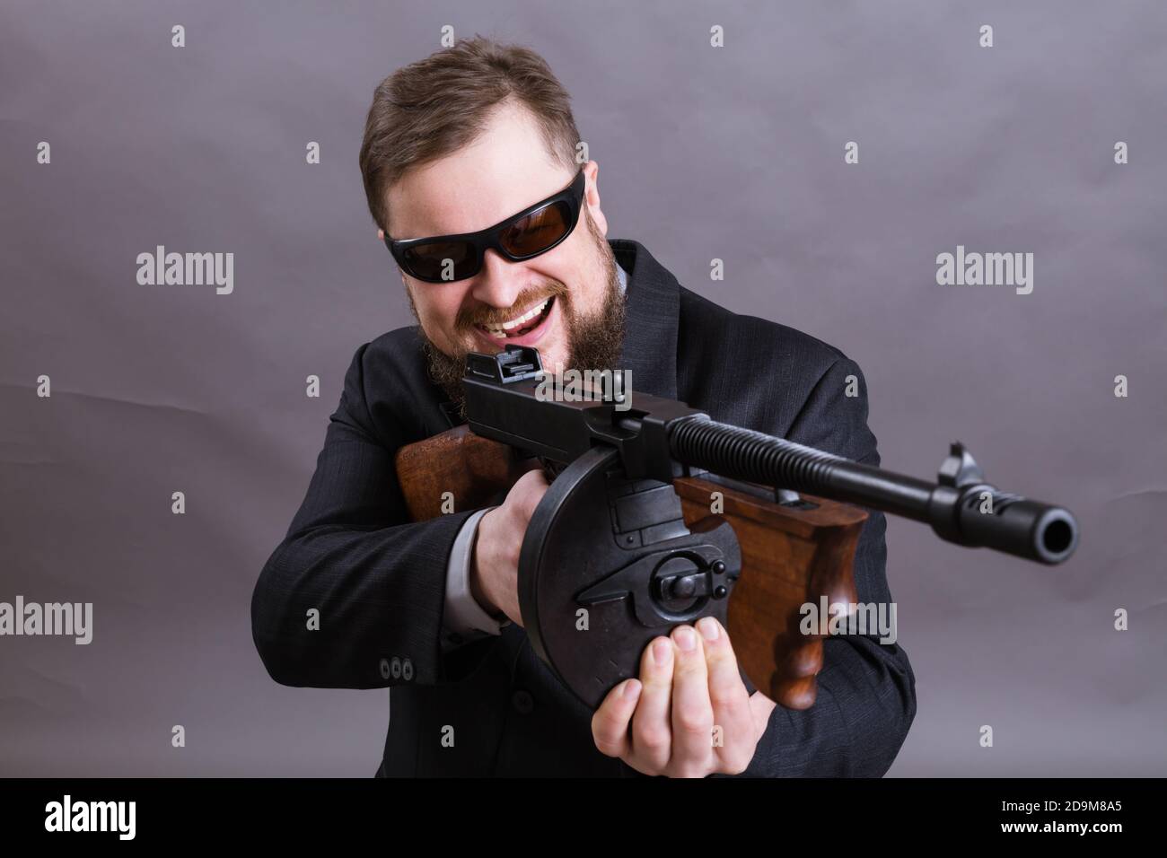 Homme à barbe mature en lunettes de soleil vêtu en costume avec tommy canon sur fond gris Banque D'Images