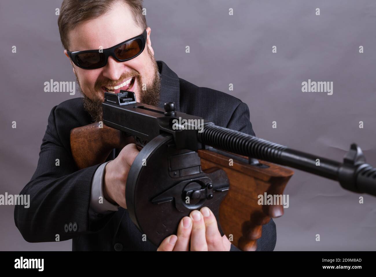 Homme à barbe mature en lunettes de soleil vêtu en costume avec tommy canon sur fond gris Banque D'Images