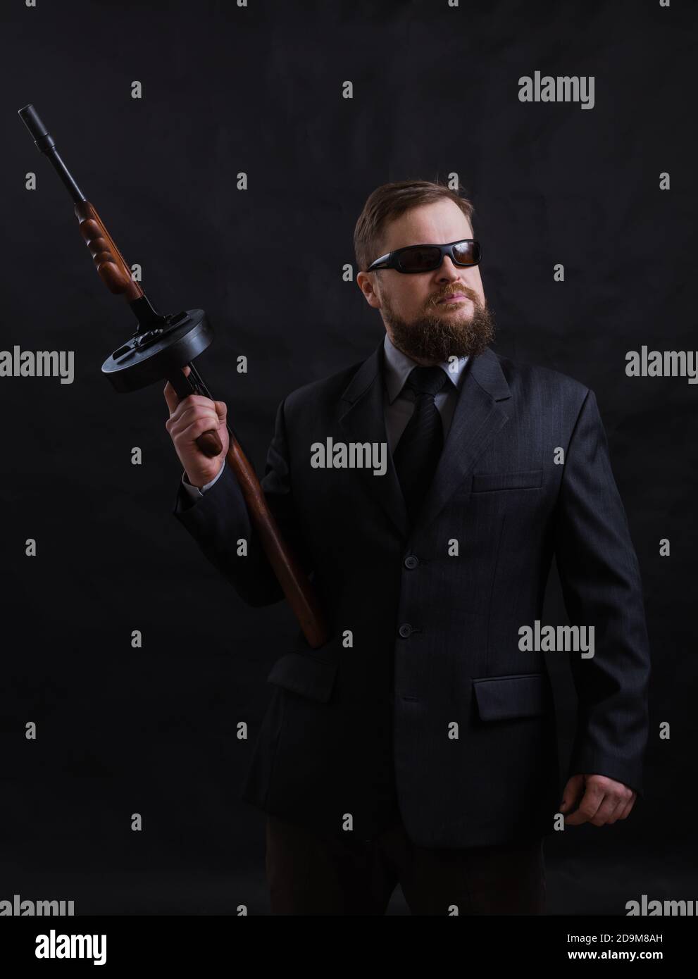Homme à barbe mature en lunettes de soleil vêtu en costume avec tommy canon sur fond noir Banque D'Images