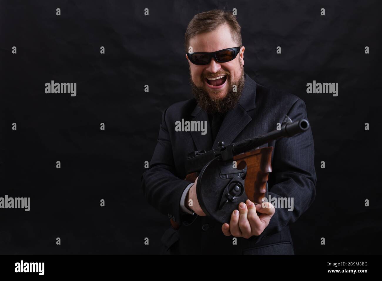 Homme à barbe mature en lunettes de soleil vêtu en costume avec tommy canon sur fond noir Banque D'Images