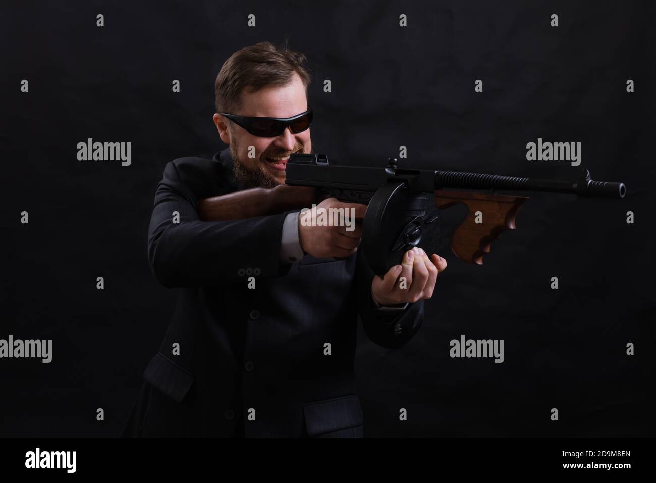 Homme à barbe mature en lunettes de soleil vêtu en costume avec tommy canon sur fond noir Banque D'Images