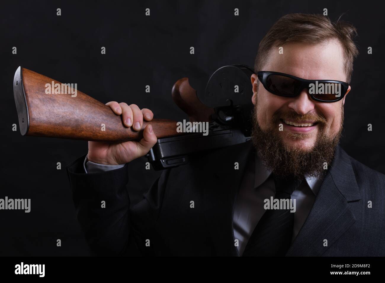 Homme à barbe mature en lunettes de soleil vêtu en costume avec tommy canon sur fond noir Banque D'Images