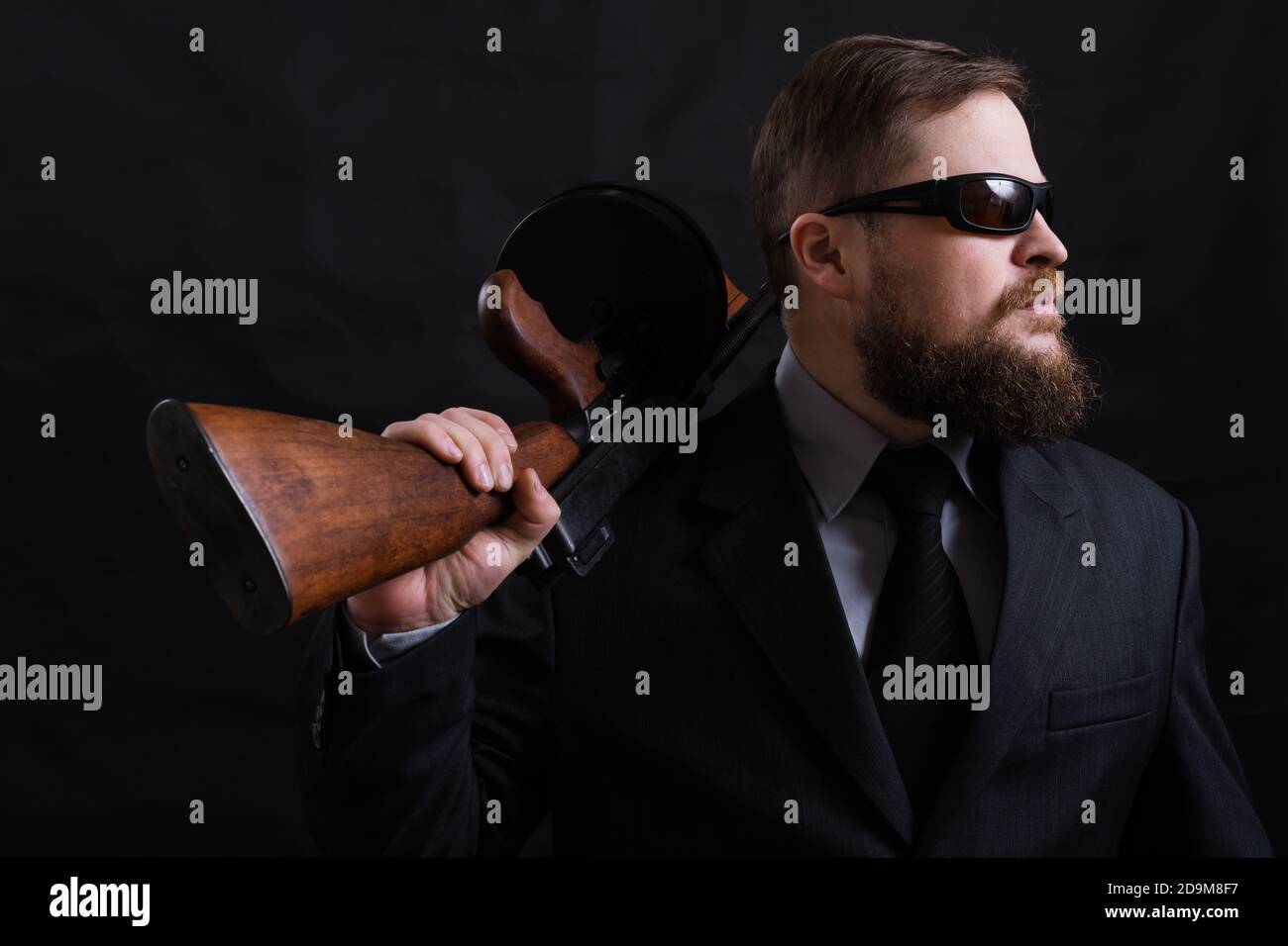 Homme à barbe mature en lunettes de soleil vêtu en costume avec tommy canon sur fond noir Banque D'Images