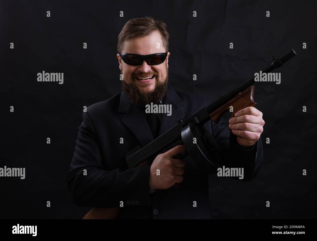 Homme à barbe mature en lunettes de soleil vêtu en costume avec tommy canon sur fond noir Banque D'Images