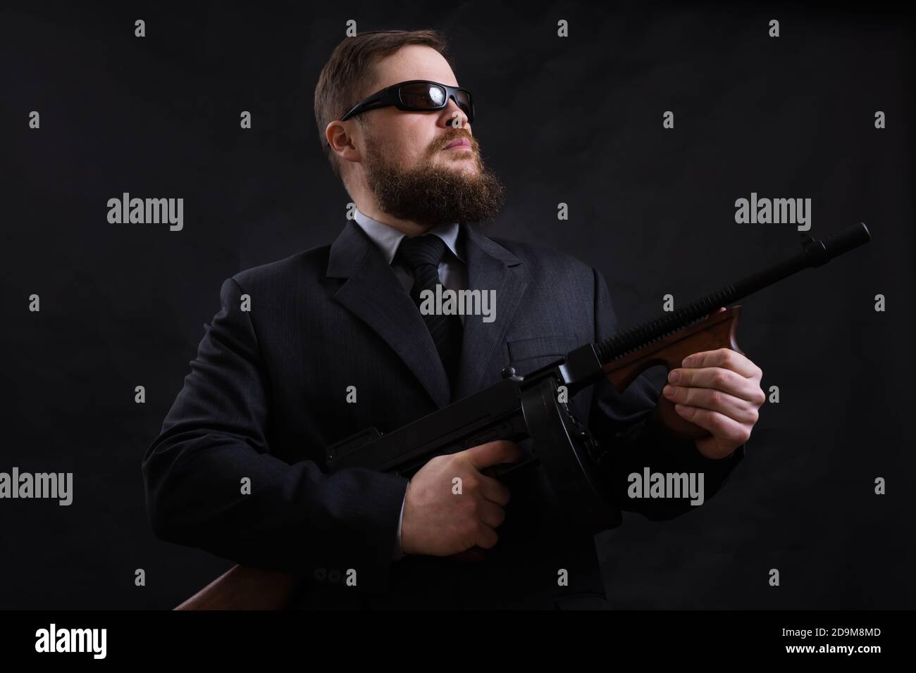 Homme à barbe mature en lunettes de soleil vêtu en costume avec tommy canon sur fond noir Banque D'Images