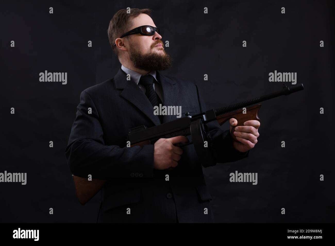 Homme à barbe mature en lunettes de soleil vêtu en costume avec tommy canon sur fond noir Banque D'Images